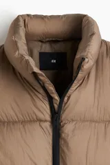 Chaleco acolchado color tostado, de nylon guateado, repelente al agua y rompevientos. Cuenta con cuello elevado, cierre bidireccional con protección para el mentón, bolsillos laterales con cierre y un bolsillo interior con botón a presión. El bajo es ajustable con cordón elástico y topes.