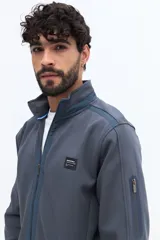 Campera negra de algodón con cierre frontal completo, cuello alto y bolsillos laterales con botón a presión. Presenta un pequeño parche rectangular con texto en el pecho izquierdo.