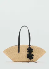 Bolso tipo shopper de fibra natural con diseño en forma de abanico. Cuenta con dos asas largas de hombro en color negro y un detalle decorativo de flores negras colgando de una de las asas.