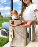 Bolso tote de cuero color taupe con textura croco y detalles en verde. Tiene doble asa de mano de cuero marrón y un dije de cuero con flecos. No tiene división interna.