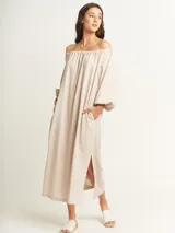 Vestido largo color beige, con escote elastizado que permite usarlo con los hombros descubiertos, mangas largas abullonadas, bolsillos laterales y tajos laterales en el ruedo.