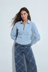 Camisa de manga larga color celeste, con cuello clásico y cierre frontal de botones. Presenta un diseño entallado con frunces estratégicos en la parte frontal que aportan textura y definen la silueta.