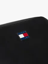 Cartera tipo bandolera de diseño compacto y minimalista, confeccionada en poliuretano con acabado liso. Presenta cierre de cremallera superior y una insignia metálica distintiva de Tommy Jeans en el centro del frente.