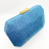 Clutch rígido rectangular forrado en lino azul, con cierre dorado en la parte superior y asa de cadena metálica dorada.