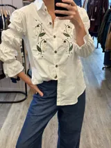 Camisa blanca de algodón con bordados florales en el frente. Presenta mangas largas con diseño abullonado, detalles de frunces y puños con volados. Cierre frontal con botones a tono.