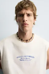 Camiseta tipo tank (sin mangas) de corte relajado, color blanco roto con efecto lavado. Presenta un estampado frontal en azul con texto en francés: "Coquillages Grillés De Côte-Marine" y "Salty Shack".