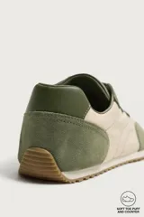 Zapatilla deportiva barefoot fabricada en combinación de piel y tejido, en tonos verde oliva y beige, con cordones elásticos amarillos y suela de goma flexible.