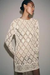 Vestido corto de punto calado en color crudo, con cuello redondo, manga larga y detalles de abalorios aplicados en forma de lentejuelas y flores.