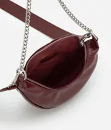 Cartera tipo riñonera de tamaño pequeño, confeccionada en poliuretano con acabado brillante. Presenta un diseño de media luna con cierre de cremallera metálica, decorado con tiradores circulares. Incluye una correa de cadena metálica para llevar al hombro o cruzada.