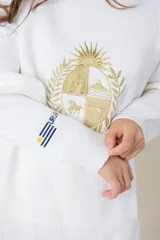 Buzo de tejido de punto color blanco, de corte holgado y cuello redondo. Presenta un bordado central en hilo dorado con el escudo nacional y un pequeño detalle bordado en la manga derecha con los colores de la bandera uruguaya.