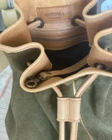 Mochila tipo bucket de gamuza marrón, con detalles de cuero liso en el tono, bolsillo frontal con cierre y cordón ajustable con tiras.