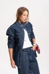 Sobrecamisa de jean oversize en color azul oscuro, con costuras a la vista en hilo blanco que resaltan su diseño. Presenta cuello clásico, cierre frontal con botones y bolsillos delanteros.