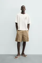 Camiseta de manga corta color blanco roto, corte relajado, con un pequeño bordado/estampado en el pecho que incluye una manzana, una rama de laurel, una trompeta y dos dados.