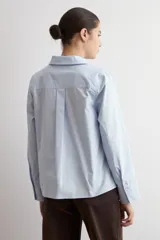 Blusa de algodón color celeste, con cuello camisero, escote en V y mangas largas con puños anchos. Presenta un diseño de corte recto y holgado.