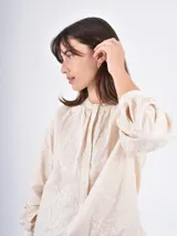 Camisa de manga larga en color crudo, confeccionada en tela con delicado bordado floral en relieve. Presenta cuello redondo con frunces, cierre frontal con botones y mangas abullonadas con puños ajustados.