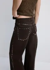 Pantalón de jean color marrón oscuro, de corte recto y tiro medio. Presenta una hilera de tachas metálicas plateadas a lo largo de la costura lateral externa.
