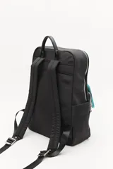 Mochila urbana de nylon en color verde militar, con detalles en cuero sintético negro. Cuenta con compartimento principal con cierre, bolsillo frontal con cierre, bolsillos laterales abiertos y asa superior. Incluye un colgante decorativo de tela y correas ajustables acolchadas.