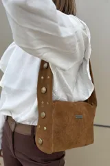 Cartera de hombro confeccionada en gamuza color habano, con diseño de correa ancha decorada con tachas metálicas circulares. Presenta cierre superior y placa metálica con logo en el frente.