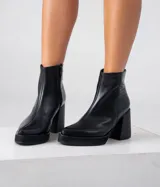 Bota corta de cuero negro con diseño combinado, presentando una textura lisa en la parte frontal y una textura tipo reptil en el talón. Cuenta con taco cuadrado alto, plataforma delantera y cierre lateral interno.