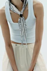 Pañuelo o bandana de algodón con estampado floral en tonos blanco, negro y azul claro. Está anudado al cuello y asegurado con un broche o piedra decorativa de color marrón oscuro y brillante.