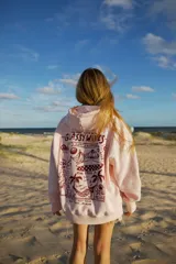 Hoodie rosa claro con estampado gráfico en la espalda en color rojo oscuro. El estampado incluye texto como "GLASSY WAVES SURF COMPANY", ilustraciones de bebidas, frutas, una tabla de surf y palmeras.