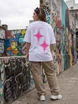 Remera oversize negra con estampado trasero en color rosa vibrante. El estampado incluye dos figuras de estrella con efecto de brillo y texto en cursiva que dice "Limits are optional", "Rush the moment" y "Own the night".