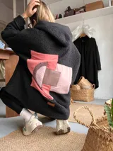 Campera de corderito negro con capucha y diseño de collage aplicado en la espalda. El collage presenta formas geométricas superpuestas en tonos rosados y marrones, confeccionado con diferentes texturas de tela.