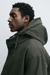 Parka color verde militar de corte relaxed fit, confeccionada en tejido de algodón con interior acolchado. Cuenta con cuello subido, capucha ajustable con cordón, cierre frontal de cremallera oculta por solapa con botones a presión y amplios bolsillos frontales de plastrón con solapa.