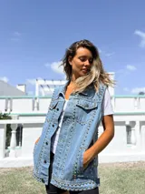 Chaleco de jean azul claro con diseño sin mangas, cuello clásico y cierre frontal con botones metálicos. Presenta una profusión de tachas plateadas distribuidas a lo largo de las costuras frontales y los bolsillos del pecho.