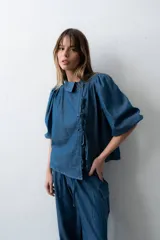 Blusa de denim liviano de corte asimétrico en tono azul, con mangas abullonadas, cuello redondo y cierre lateral con lazos para atar y botones.