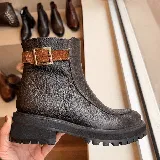 Bota de cuero texturizado en color marrón oscuro, con suela track de goma negra. Presenta una tira decorativa de gamuza en tono marrón más claro con una hebilla metálica envejecida en el lateral.