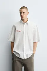 Camisa de algodón blanca con finas rayas verticales negras, corte relajado (relaxed fit) y manga corta. Presenta un bordado en hilo rojo en el lado izquierdo del pecho.