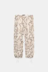 Pantalón bombacho de tiro medio con estampado de piel de serpiente en tonos beige y marrón. El bajo es abullonado y ajustable con lazada. Presenta detalle de hilo metalizado a contraste y cierre lateral con cremallera oculta.