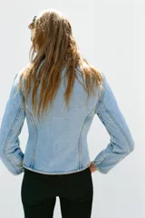 Chaqueta denim entallada de cuello redondo y manga larga, con cierre frontal de cremallera metálica. Presenta detalles de aplicaciones en el mismo tejido y pasamanería frontal de inspiración militar.
