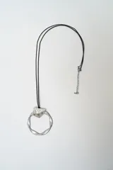 Collar con cordón negro y dije circular de metal plateado con textura irregular y pieza geométrica superior.