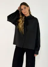 Camisa negra de corte holgado con cuello tipo bebé con volados y lazo de ajuste, cierre frontal con botones y mangas largas abullonadas con puños abotonados.