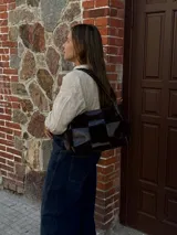 Cartera de hombro confeccionada en cuero y gamuza con diseño de damero, combinando paneles de texturas lisas y aterciopeladas. Presenta una manija trenzada y detalles de costuras artesanales en los laterales.