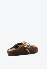 Zueco de gamuza marrón, estilo slip-on, con cordones frontales decorativos, abalorios y suela plana.