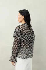 Camisa blanca de encaje bordado con diseño semitransparente, silueta fluida y mangas largas. Se destaca por los volados en el frente y hombros.