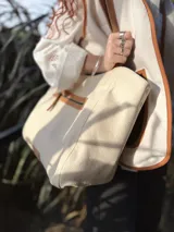 Bolso tipo tote color crema, con detalles en cuero color marrón. Tiene un bolsillo con cierre en la parte superior y un parche de cuero con el logo de la marca en la parte inferior.