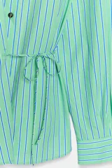 Camisa de manga larga con cuello solapa, cierre frontal con botones y diseño cruzado con lazada lateral en el mismo tejido. Presenta un estampado de rayas verticales en tonos verdes y azules.