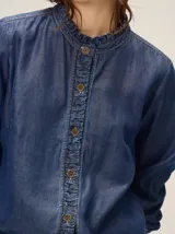 Camisa de denim liviano color azul, con cuello mao y detalles de volados fruncidos en el cuello y la botonera frontal. Presenta mangas largas con puños rectos y corte relajado.