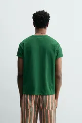 Camiseta de corte relajado, color verde, con cuello redondo y manga corta. Presenta un texto bordado en relieve con letras amarillas y contorno oscuro en el centro del pecho.