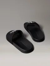 Ojotas tipo slide Calvin Klein Jeans, color negro, con banda superior de lona que presenta el logo de la marca bordado en blanco. Posee suela de EVA con plantilla ergonómica moldeada y el nombre de la marca grabado en el lateral.
