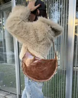 Cartera de cuero vacuno color marrón con diseño semicircular, detalle de argollas metálicas en el borde inferior y correa larga ajustable.