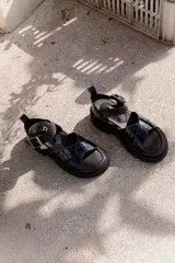 Sandalias negras de estilo chunky con tiras cruzadas en charol y base de goma dentada. Cuentan con una tira superior desmontable y cierre con hebilla metálica en el tobillo.