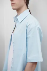 Camisa de corte oversize y diseño unisex, color celeste, con cuello clásico, manga corta amplia y bolsillo frontal. En la espalda presenta un canesú con pliegue central que aporta amplitud y caída fluida.