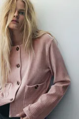 Chaqueta corta de velour acanalado color rosado, con cuello redondo, cierre frontal de botones marrones y dos bolsillos de parche con botón en la parte delantera. Presenta hombros caídos y mangas largas.