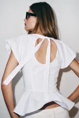 Top de popelina blanca con cuello redondo, mangas cortas abullonadas y detalle de abertura en la espalda con lazada. Presenta bajo en evasé tipo peplum y cierre lateral con cremallera oculta.