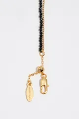 Pulsera doble dorada que combina una cadena de eslabones con una tira de cuentas negras de cristal. Cuenta con un dije colgante en forma de corazón esmaltado y un dije de perla barroca.
